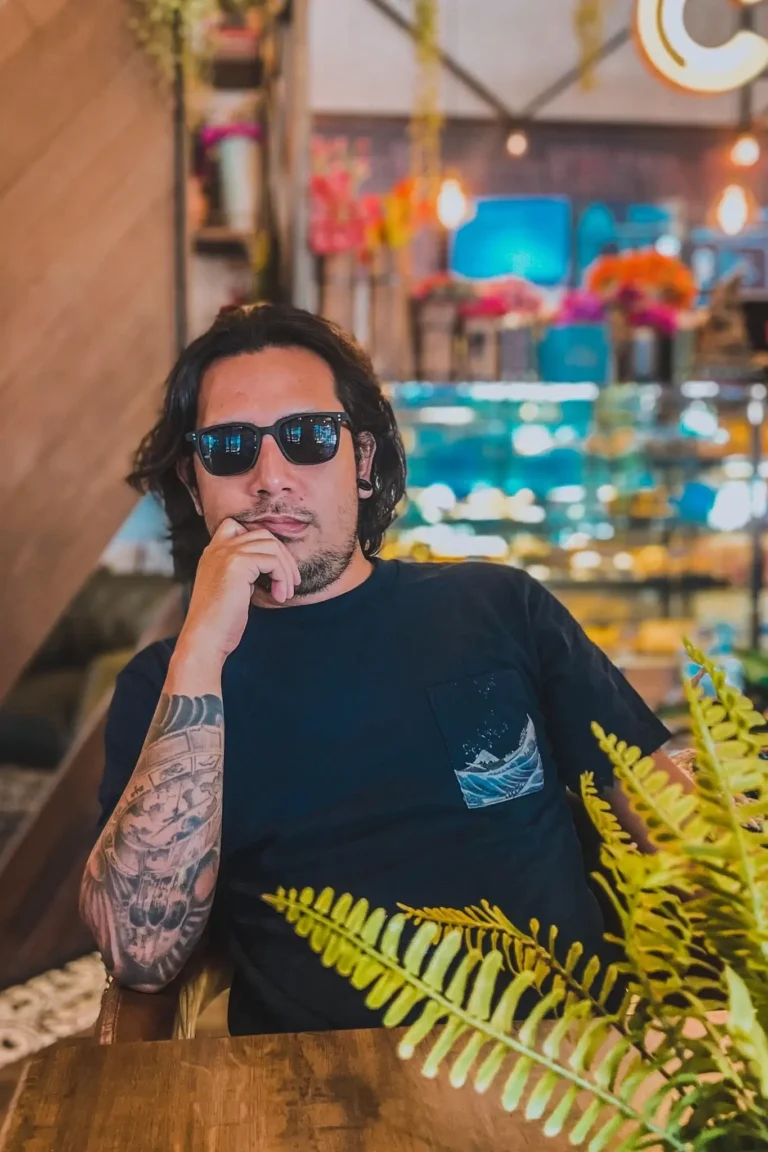 Portrait of Noel in casual attire, showcasing his tattoos and sunglasses in a cozy cafe setting.